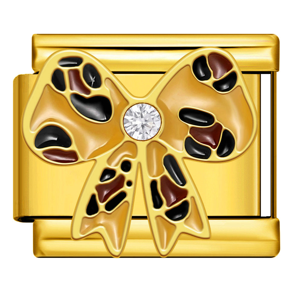 A gold-toned bow-shaped charm or brooch with black and dark brown enamel sections, a clear round gemstone at the center, and a polished metallic finish on a plain background.